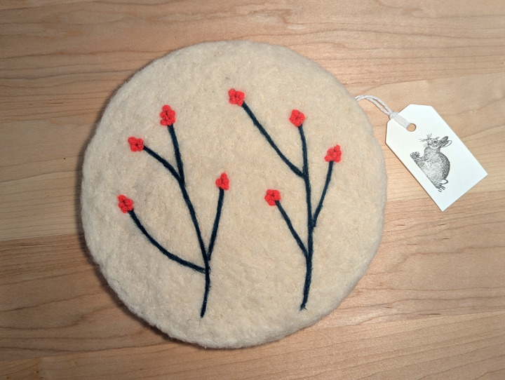 A felted circular white trivet with a stylized floral design in green and pink.