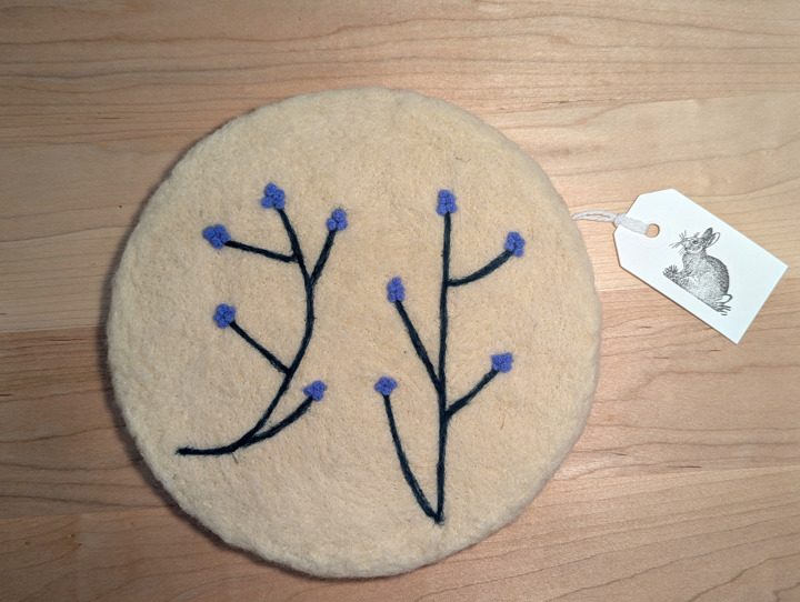 A felted circular white trivet with a stylized floral design in green and periwinkle.