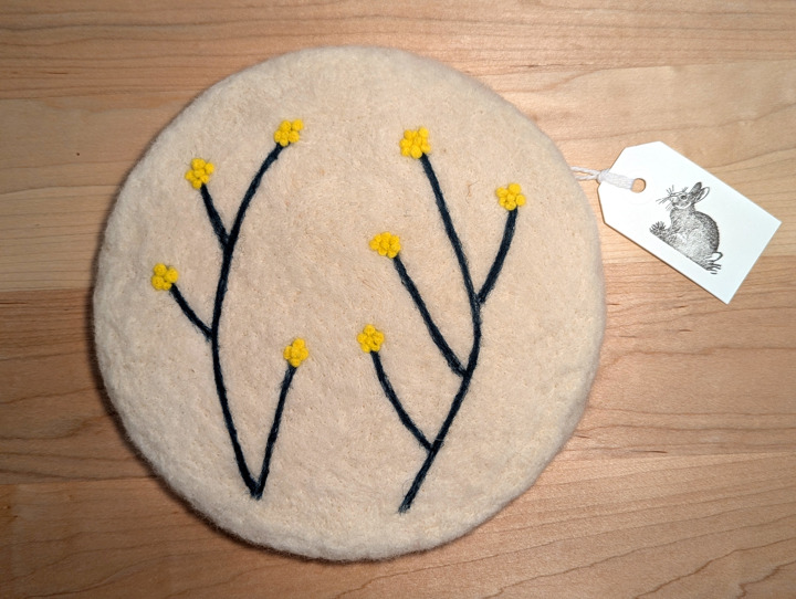 A felted circular white trivet with a stylized floral design in green and yellow.