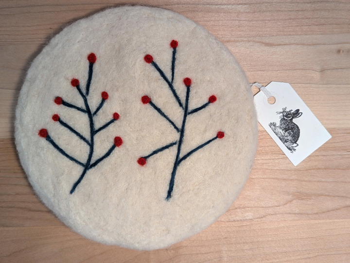 A felted circular white trivet with a stylized floral design in green and red.