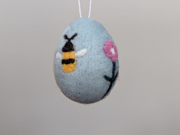 A felted sky blue egg decorated with stylized flowers and a happy bee.