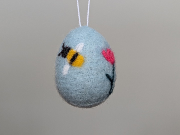 A felted sky blue egg decorated with stylized flowers and a happy bee.