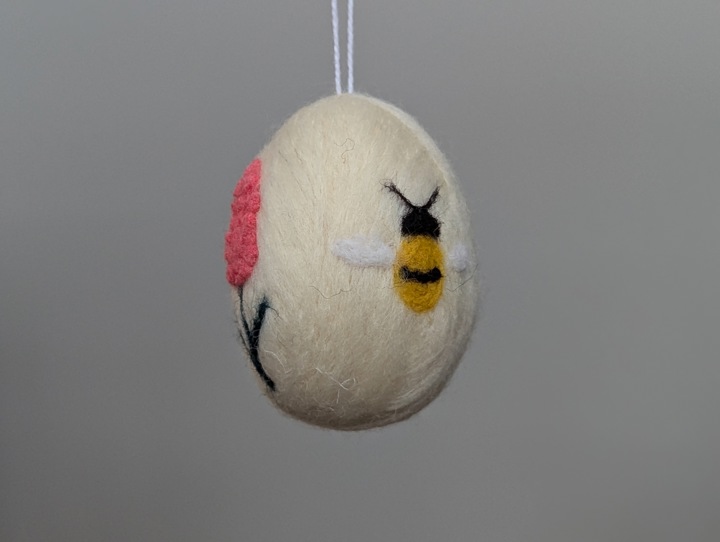 A felted white egg decorated with stylized flowers and a happy bee.