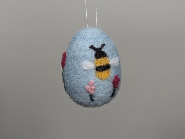 A small felted sky blue egg decorated with stylized flowers and a happy bee.