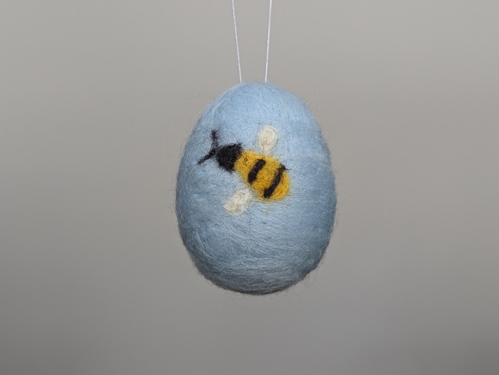 A small felted sky blue egg decorated with stylized flowers and a happy bee.