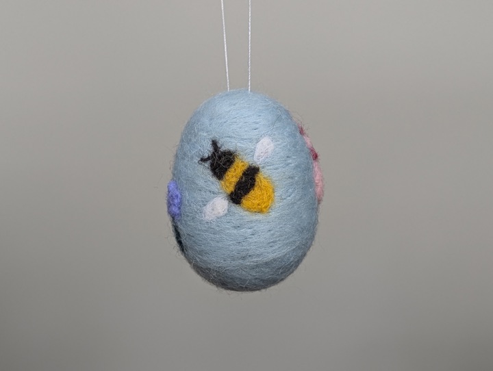 A small felted sky blue egg decorated with stylized flowers and a happy bee.