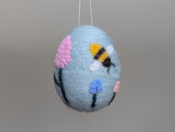 A felted sky blue egg decorated with stylized flowers and a happy bee.