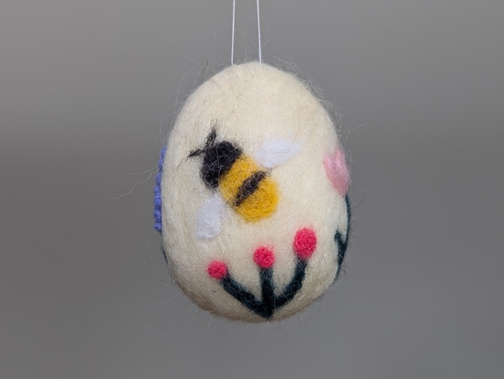 A felted white egg decorated with stylized flowers and a happy bee.