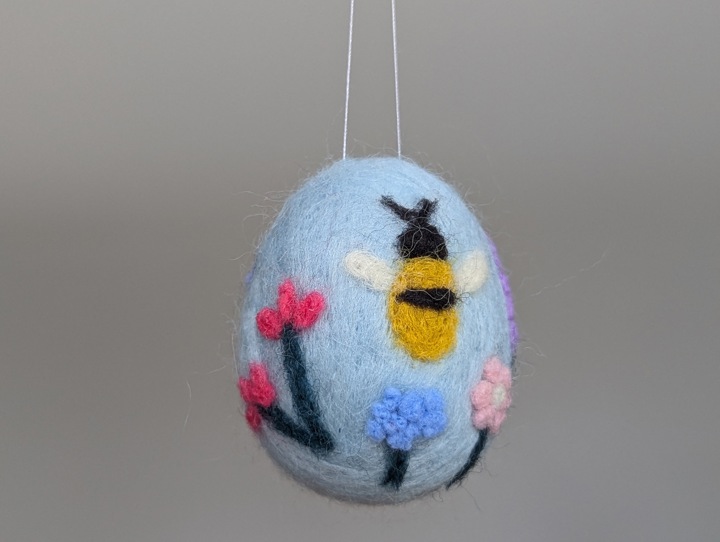 A felted sky blue egg decorated with stylized flowers and a happy bee.