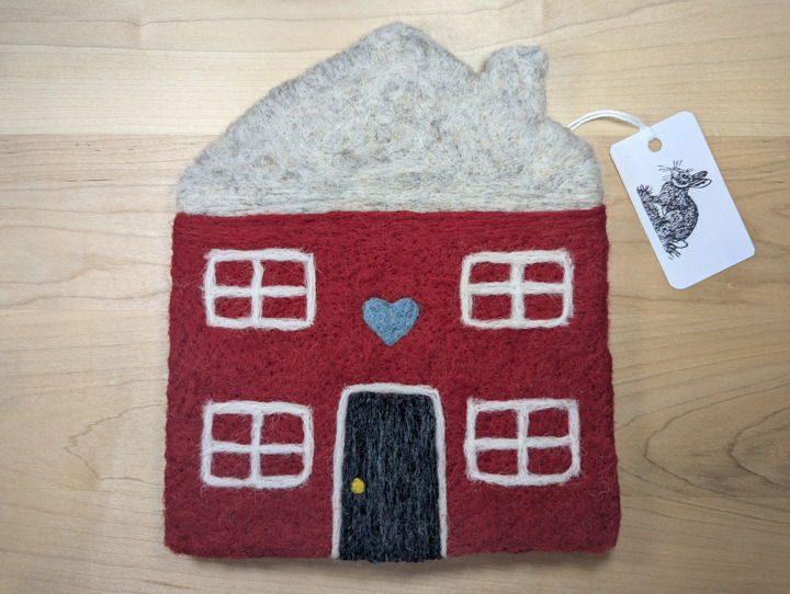 A felted red house with a light grey roof and a dark grey door with a country blue heart motif above it.
